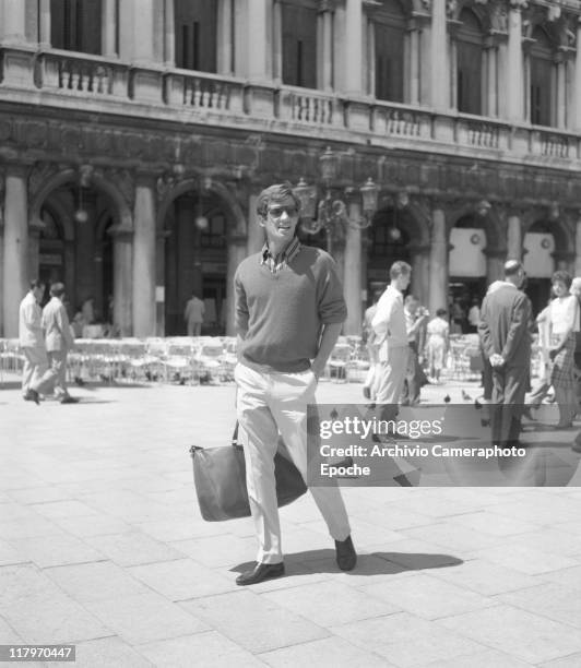 French actor Jean-Paul Belmondo, wearing a striped shirt, a sweater and sunglasses, holding a bag and standing in St Mark's Square, Venice, 1960.