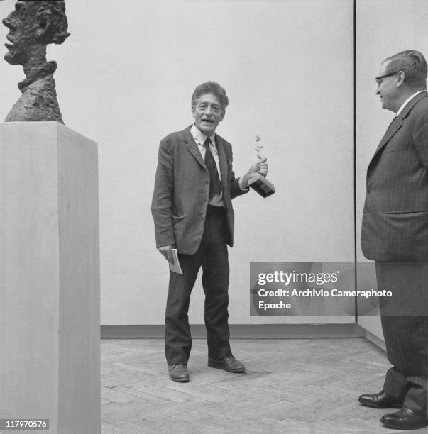 Swiss sculptor Alberto Giacometti, wearing a blazer and a tie, standing in the exhibition room next to the sculpture 'La Grande Tete De Diego',...