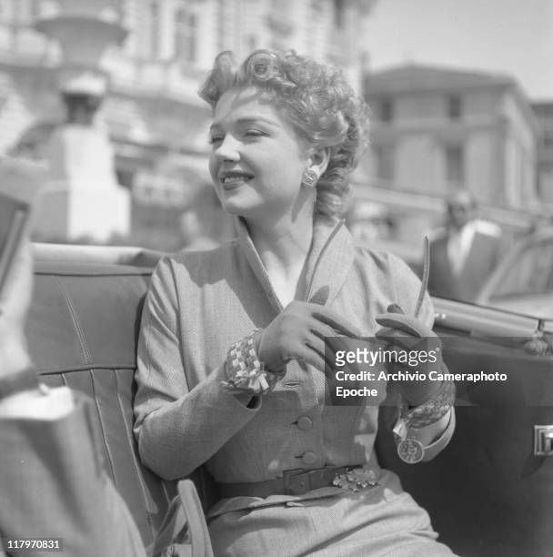 American actress Anne Baxter, wearing a tailleur, a belt and gloves, holding sunglasses and looking around, while sitting in a car, Cannes 1953.