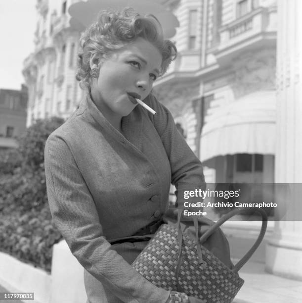 American actress Anne Baxter, wearing a tailleur ang gloves, a cigarette in her mouth, looking for something in her handbag, Cannes, 1953.