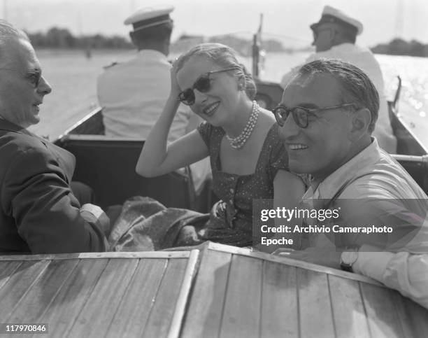 American actress Joan Fontaine wearing a fancy dress, a pearl necklace and sunglasses, sitting on a water taxi with two men, Torcello island ahead,...