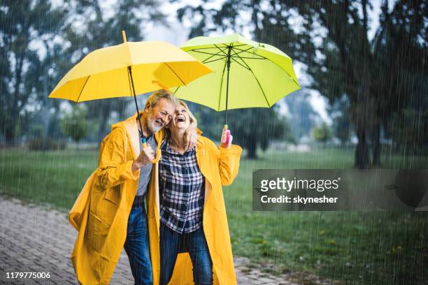 glückliches reifes paar in gelben regenmänteln, die an einem regnerischen tag unter regenschirmen spazieren gehen. - regenschirm stock-fotos und bilder