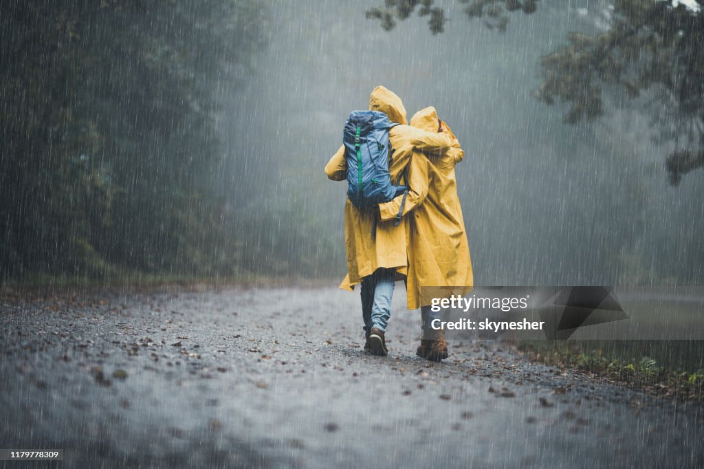 Back view of embraced couple in raincoats hiking on a rain.