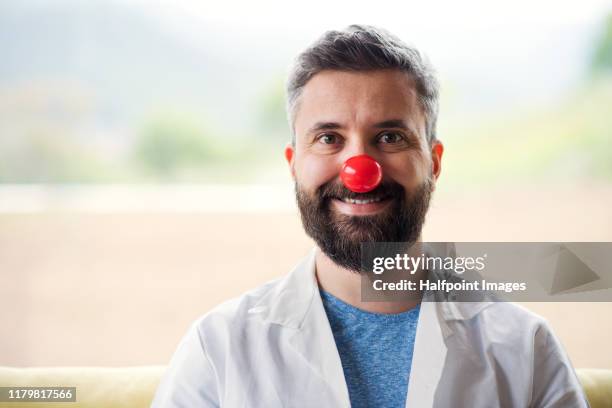 a front view portrait of healthcare worker or doctor with red clown nose standing indoors. - clown's nose stock pictures, royalty-free photos & images