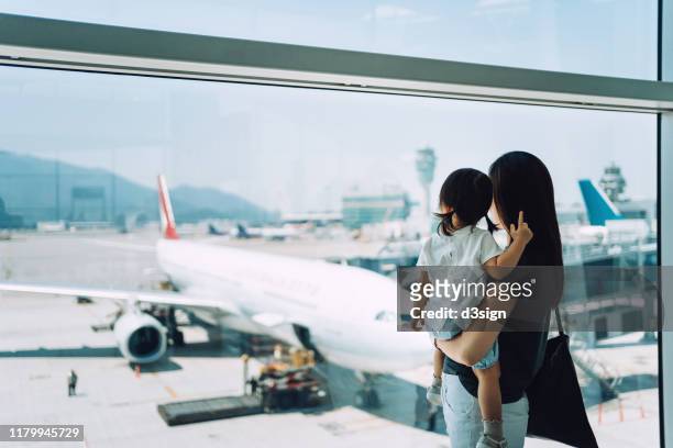 joyful young asian mother embracing cute little daughter looking at airplane through window at the airport while waiting for departure - baby gate imagens e fotografias de stock