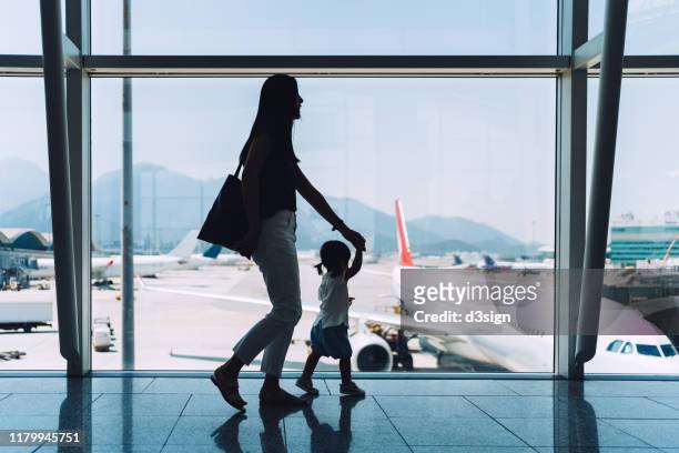 silhouette of joyful young asian mother holding hands of cute little daughter looking at airplane through window at the airport while waiting for departure - airport family stock-fotos und bilder