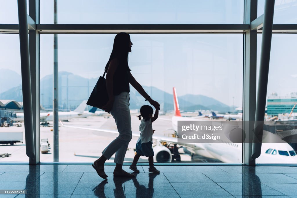Silhouette of joyful young Asian mother holding hands of cute little daughter looking at airplane through window at the airport while waiting for departure