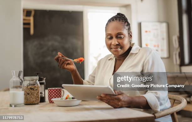 smiling african american woman using a tablet and eating breakfast - mature women eating stock pictures, royalty-free photos & images