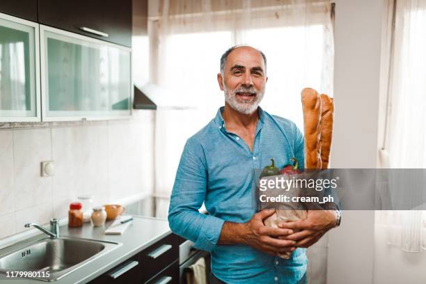 mature man holding bag with groceries in the kitchen - baguette white stock pictures, royalty-free photos & images