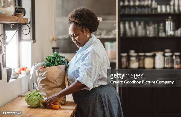 lächelnde afroamerikanische frau auspacken lebensmittel in ihrer küche - african woman shopping stock-fotos und bilder