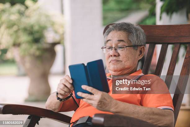 een aziatische chinese senior man ontspannen op zijn bank in de voorkant van zijn huis te wachten op zijn familie terug naar huis voor chinees oudejaarsavond reünie diner tijdens het gebruik van zijn telefoon voor communicatie - 75 year old pensioners stockfoto's en -beelden