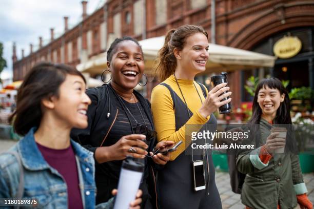 happy female friends with drinks walking in city - multikulturelle gruppe stock-fotos und bilder