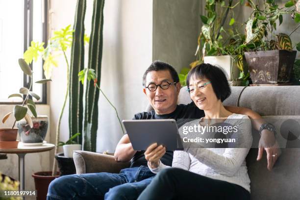 asiatische spaar mit einem digitalen tablet auf dem sofa - older asian couple stock-fotos und bilder