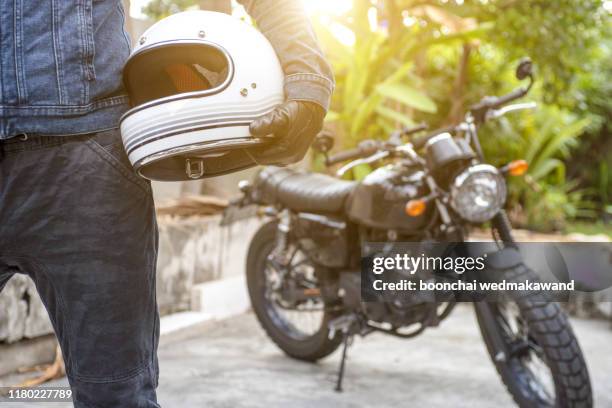 handsome man in jean is holding a helmet and vintage motorcycle blur background - vintage motorcycle stockfoto's en -beelden