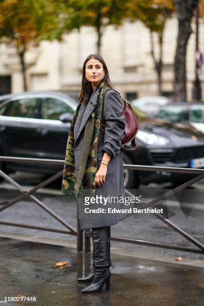 Julia Haghjoo wears earrings, bracelets, a khaki and yellow camouflage scarf, a grey oversized coat with shoulder pads, a burgundy studded bag, black...