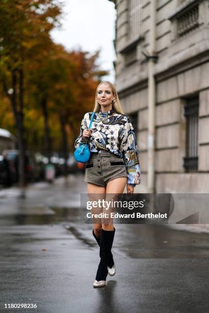 Leonie Hanne wears earrings, a necklace, a white long sleeves hi-neck blouse with grey swan and mermaids print, brown Prince of Wales shorts, a blue...