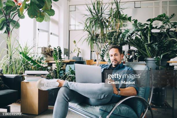 portrait of confident creative businessman with mobile phone and laptop sitting on seat in office - sitting and using smartphone studio stockfoto's en -beelden