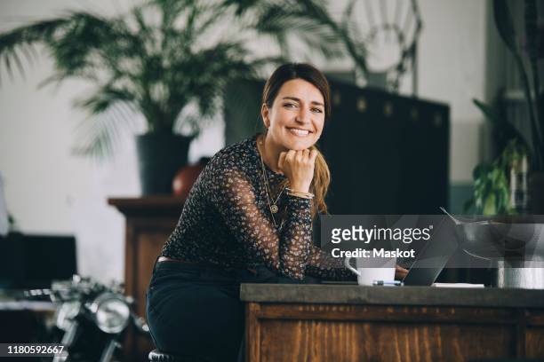 portrait of smiling businesswoman with hand on chin sitting at kitchen island in creative office - woman portrait kitchen laptop stock-fotos und bilder