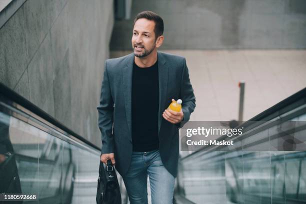 confident businessman with drink looking away while moving on escalator in subway - casual business stock-fotos und bilder