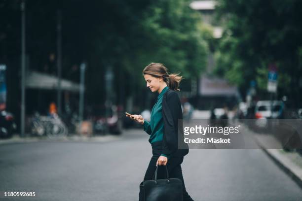 confident businesswoman using smart phone while crossing street in city - seitenansicht stock-fotos und bilder