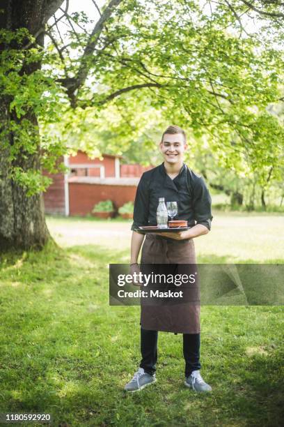 full length portrait of smiling confident young waiter standing on grass at outdoor cafe - kellner tablett stock-fotos und bilder
