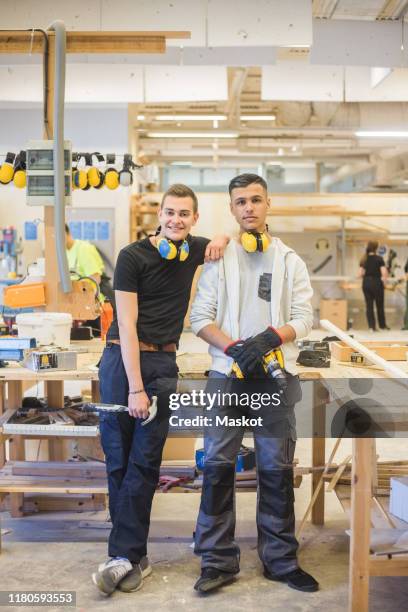 full length of smiling young male trainee leaning on coworker's shoulder while holding work tools at workshop - ausbildung und tischler stock-fotos und bilder