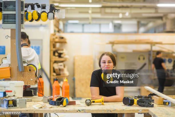 portrait smiling young female carpentry trainee leaning on workbench with power tools at illuminated workshop - deeltijdbaan stockfoto's en -beelden