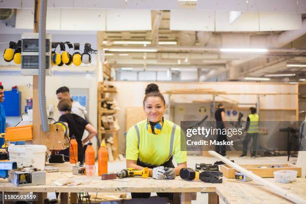 portrait of smiling young female trainee leaning on workbench with power tools at illuminated workshop - female builder stock pictures, royalty-free photos & images