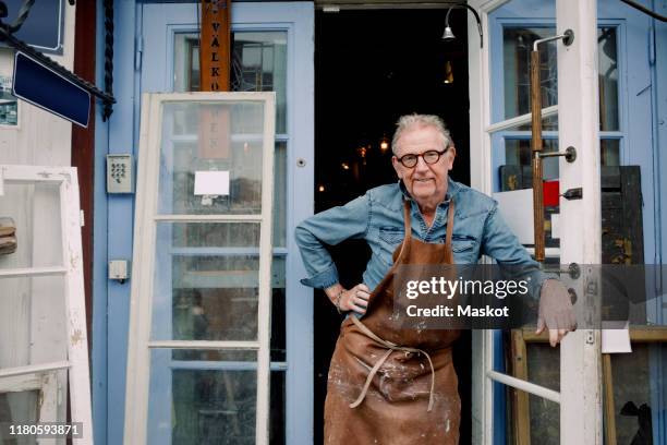 portrait of senior hardware store owner standing with hand on hip while leaning on door - menselijke leeftijd stockfoto's en -beelden