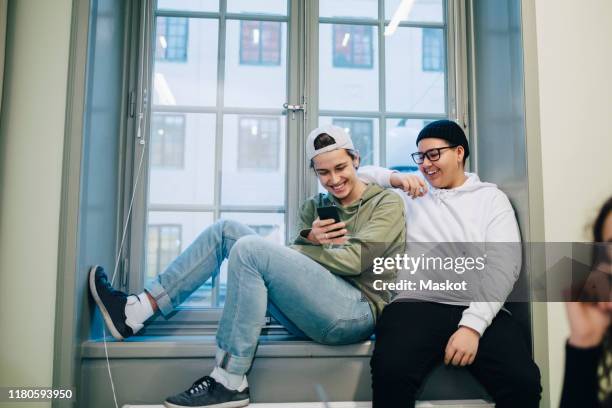 smiling male teenage students using phone while sitting on window in classroom - sekundarschule stock-fotos und bilder