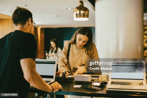 man looking at woman signing at reception desk in hotel lobby - receptionist fotografías e imágenes de stock