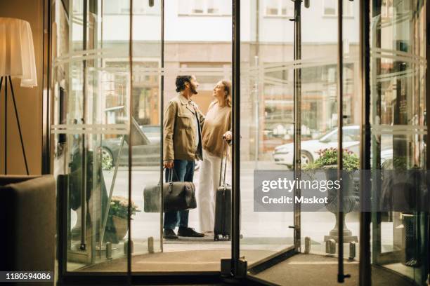 couple with luggage talking while standing at doorway of hotel seen through glass - hotel stadt stock-fotos und bilder