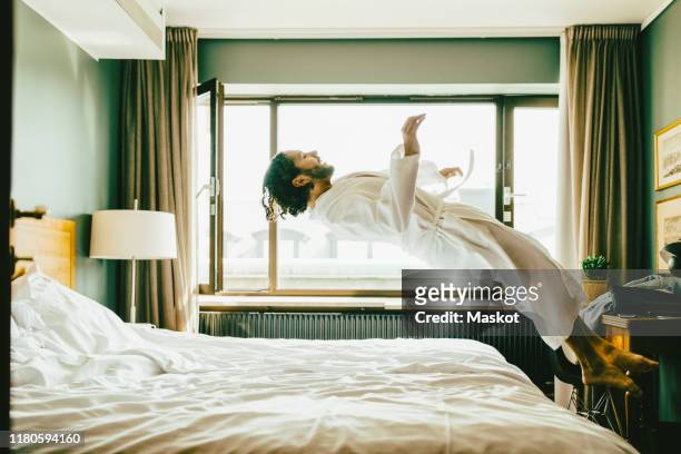 playful man wearing robe jumping on bed at hotel - hotel stock-fotos und bilder