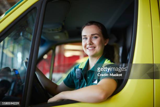 portrait of smiling young female paramedic driving ambulance in parking lot - paramedics stock-fotos und bilder