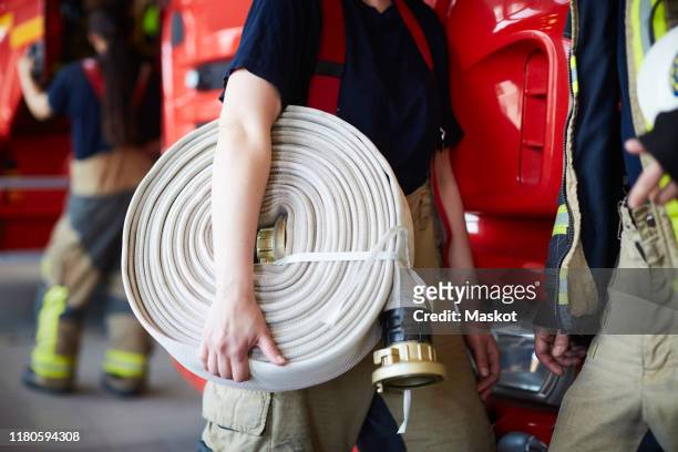 midsection of female firefighter holding rolled up fire hose while standing with coworker at fire station - brandslang stockfoto's en -beelden