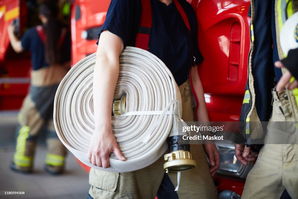 Midsection of female firefighter holding rolled up fire hose while standing with coworker at fire station