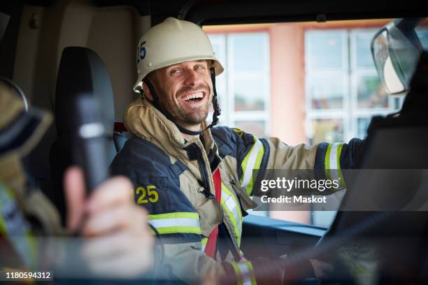 smiling firefighter looking at coworker while sitting in fire engine - fireman uniform stock pictures, royalty-free photos & images