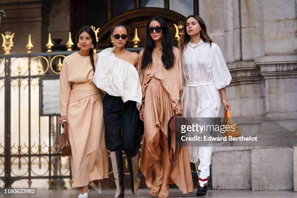 Bettina Looney wears earrings, a beige long dress, a brown bag, white pointy pumps ; Anna Rosa VItiello wears sunglasses, a gathered flowing...