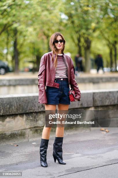 Gala Gonzalez wears sunglasses, a burgundy leather oversized jacket, a burgundy and white striped top, navy blue denim shorts, a red studded and...