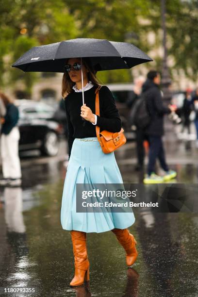 Guest wears a dark pullover, a white shirt with ruffled collar, a pale blue pleated skirt, orange leather boots, a brown leather bag, holds an...