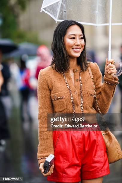 Aimee Song wears a brown wool jacket, red leather shorts, a light brown quilted bag, holds an umbrella under the rain, outside Chanel, during Paris...