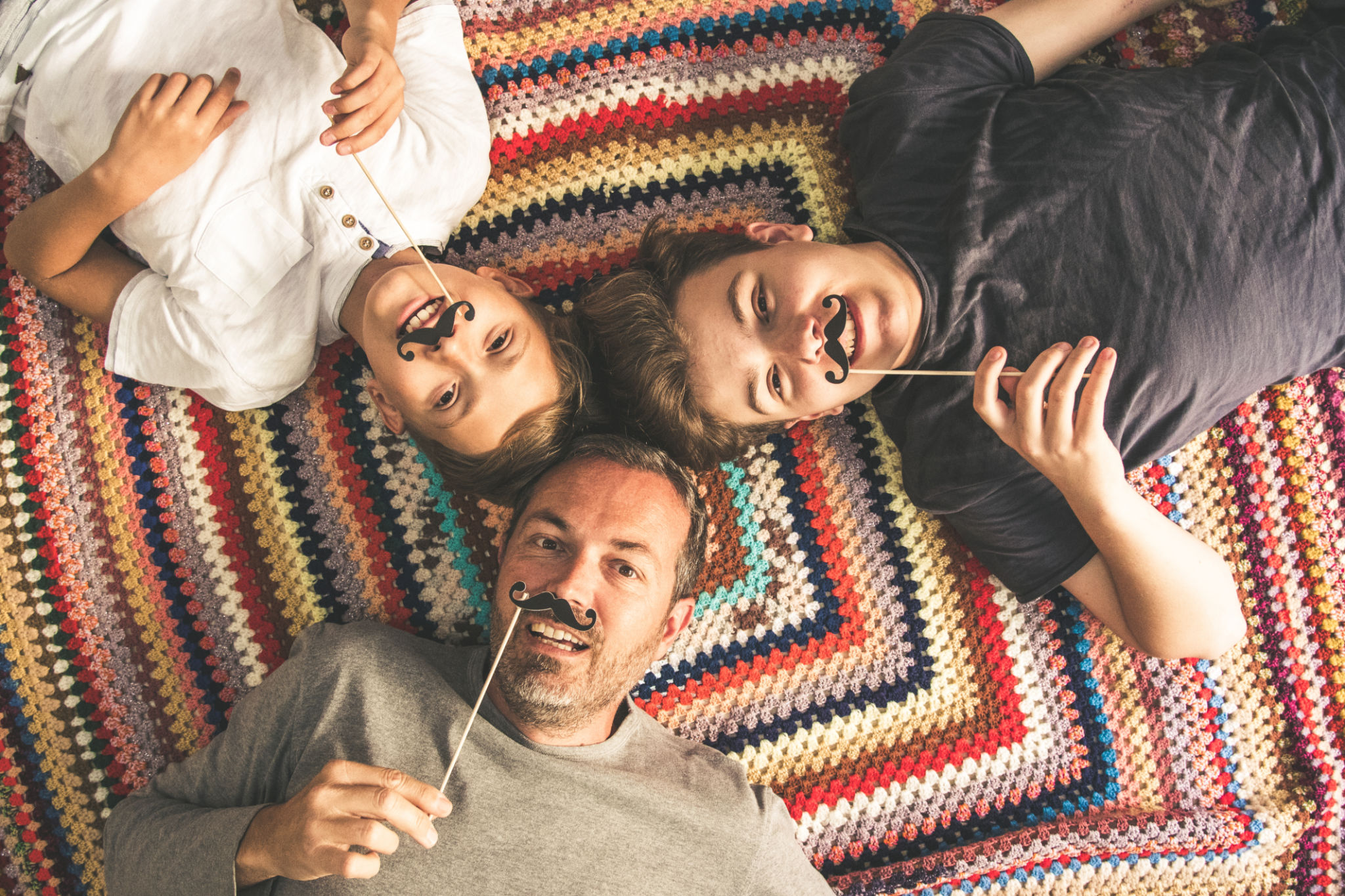 Father and two sons enjoying together lying on a colorful blanket. Tree men of different ages smiling playing with fake mustache. Top view of a couple of teen and their dad. Father and two sons enjoying together lying on a colorful blanket. Tree men of different ages smiling playing with fake mustache. Top view of a couple of teen and their dad.