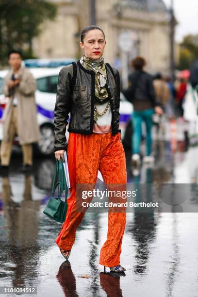 Guest wears a scarf, a black trimmed bronze-tone jacket, orange veil pants with velvet arabesque pattern, a forest-green bag, colorful print pointy...