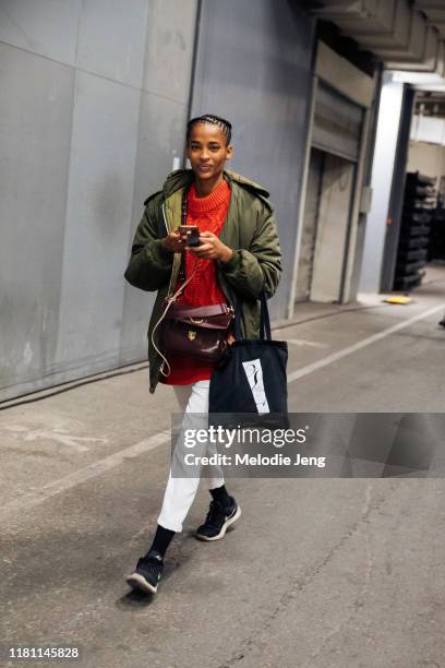 Model Ana Flavia wears a green jacket, red knit sweater, brown purse, white pants, black Nike sneakers, and checks her phone after the Off-White show...