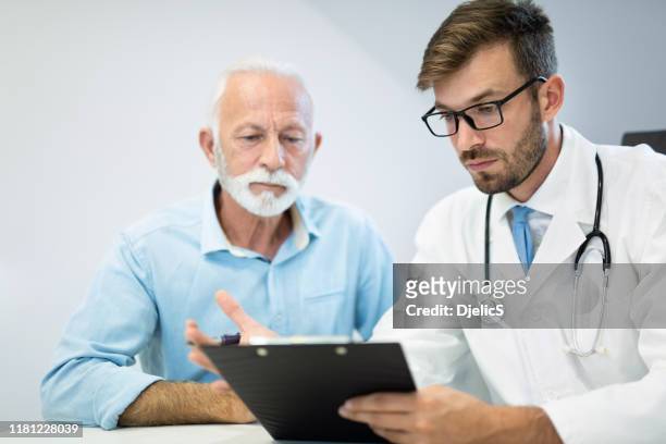 junger arzt und männlicher seniorpatient, der sich eine krankenakte ansieht. - doctor with male patient reading notes stock-fotos und bilder
