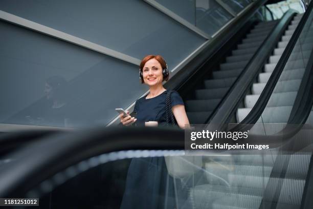 young businesswoman with coffee and smartphone indoors on escalator in city, listening to music. - escalator photos et images de collection