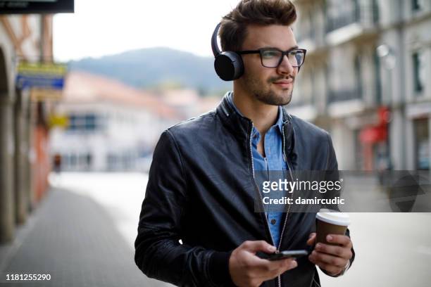 glimlachend jong man luisteren naar muziek en het drinken van koffie buitenshuis - young man listening to music on smart phone outdoors stockfoto's en -beelden