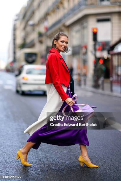 Landiana Cerciu wears a lustrous purple flowing dress, a chain belt, yellow heels, a red and white coat, a pale pink and light purple bag, outside...