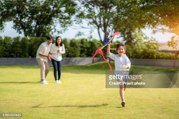 asian daughter running to mother and father, she enjoying the play kite  with her family in park at village. parenthood and childhood lifestyle or love and bonding in concept. - thailand kite stock-fotos und bilder