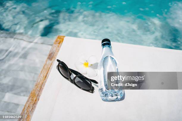 water bottle, sunglasses and flower petal on a deck chair by the swimming pool under sunshine, summer time fun, essentials, tourism and vacation concept - beach deck chairs stock-fotos und bilder